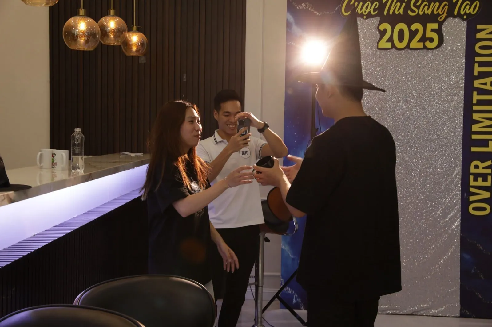 Thí sinh và nhân sự Ethan trò chuyện, quay hình bên quầy bar trước backdrop Cuộc Thi Sáng Tạo 2025 ở hậu trường Over Limitation